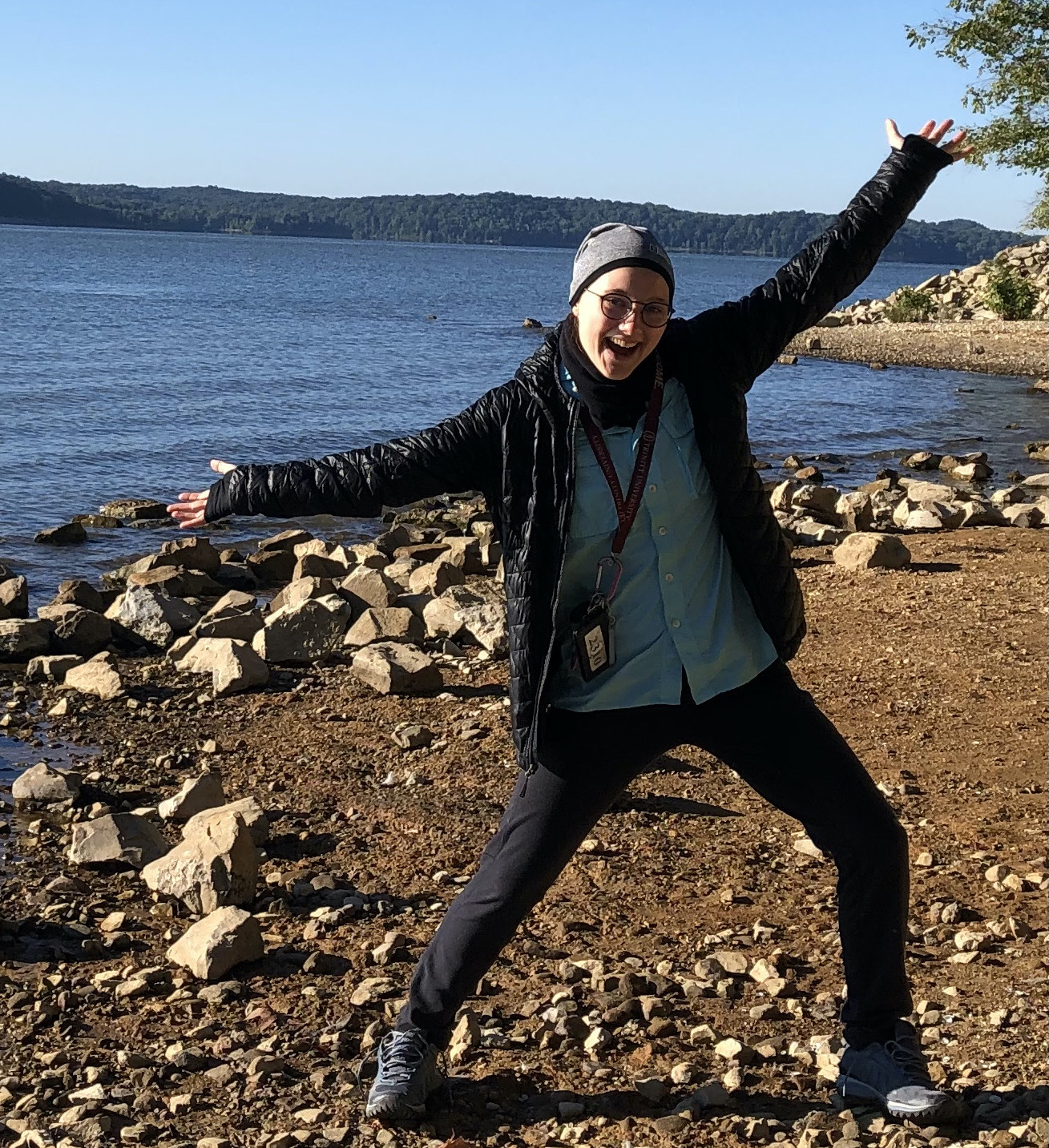 Photo of a young white woman standing at the shore of a lake with arms outstretched and an excited face